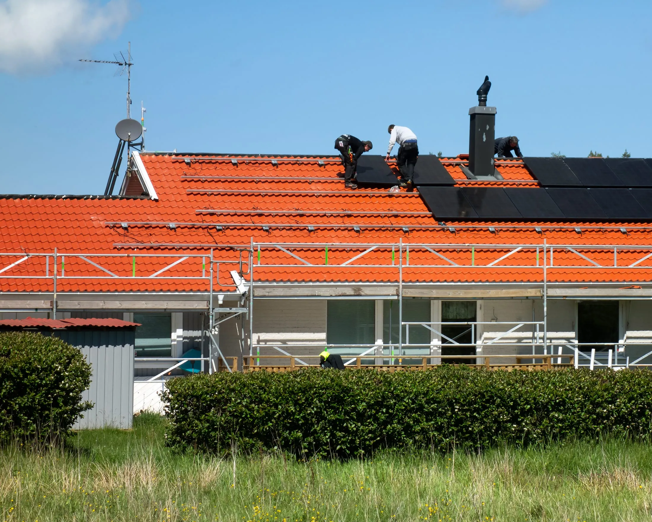 Three installers laying black solar panels across a terracotta tiled roof, working from scaffolding.